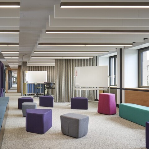 New Work Workshopbereich mit Baffelleuchten Workshop-Büroraum mit bunten Polstermöbeln und Sitzwürfeln, darüber sind in zwei Reihen hintereinander mehrere lineare Akustikbaffeln und Leuchten zu sehen.