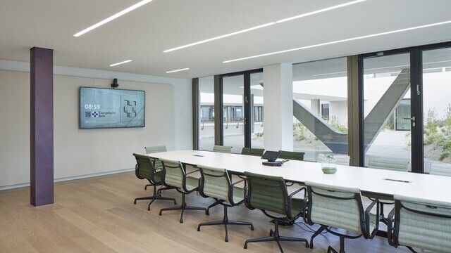 EVB conference room with an oval meeting table and office chairs around it in the centre, windows around the outside provide a view of the outside, linear recessed luminaires (PALADIO) are visible in the white ceiling.