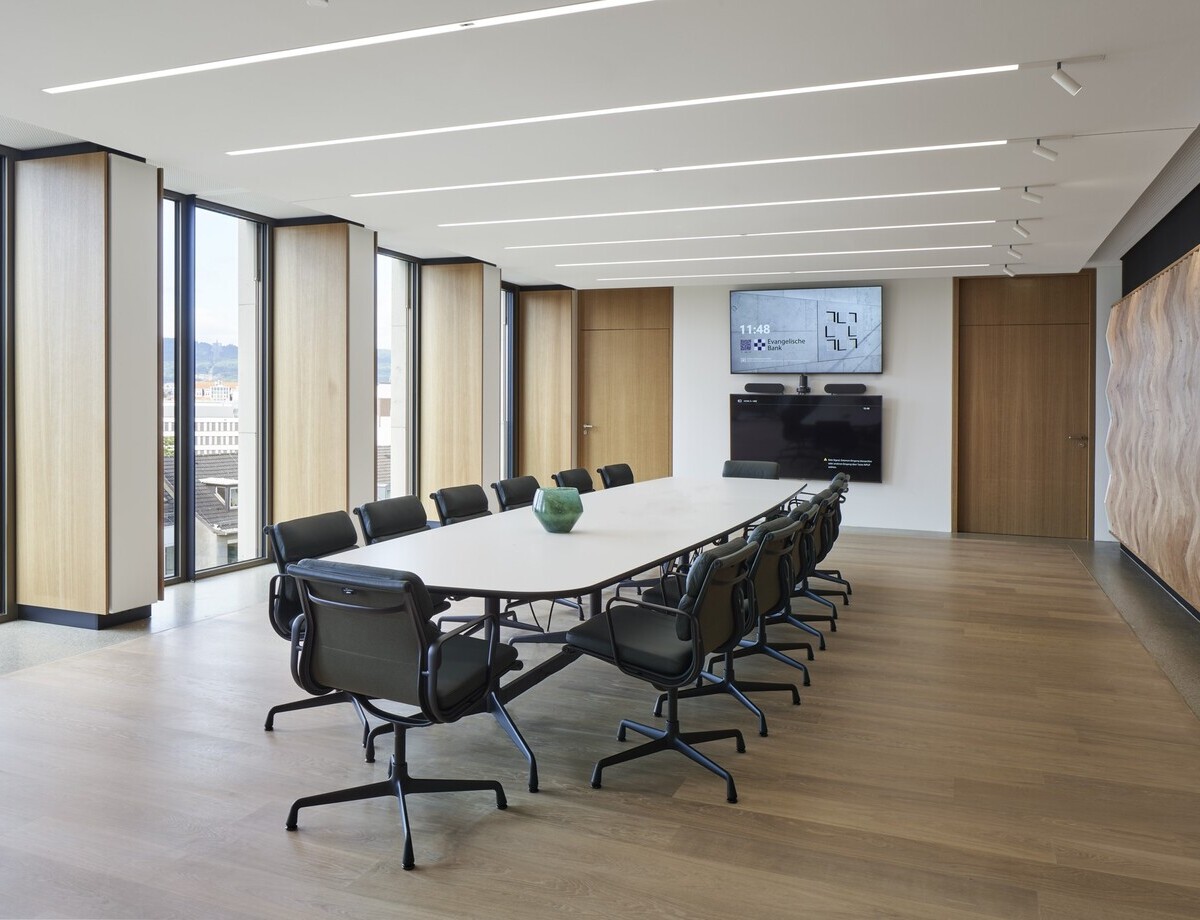 Konferenzraum der EVB mit einem ovalen Meetingtisch und Bürostühlen darum herum in der Mitte, außenherum geben Fenster den Blick nach draußen frei, in der weißen Decke sind lineare Einbauleuchten (PALADIO) sichtbar, zu einer Wandseite sind am Ende der Leuchten noch zylindrische Spots (EVOLUTION SPOT) anschließend eingebaut.