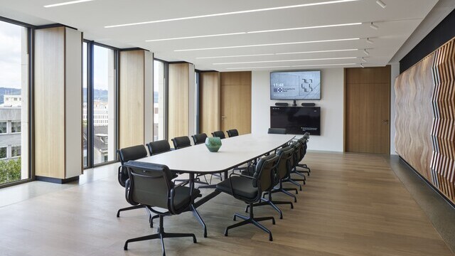Konferenzraum der EVB mit einem ovalen Meetingtisch und Bürostühlen darum herum in der Mitte, außenherum geben Fenster den Blick nach draußen frei, in der weißen Decke sind lineare Einbauleuchten (PALADIO) sichtbar, zu einer Wandseite sind am Ende der Leuchten noch zylindrische Spots (EVOLUTION SPOT) anschließend eingebaut.