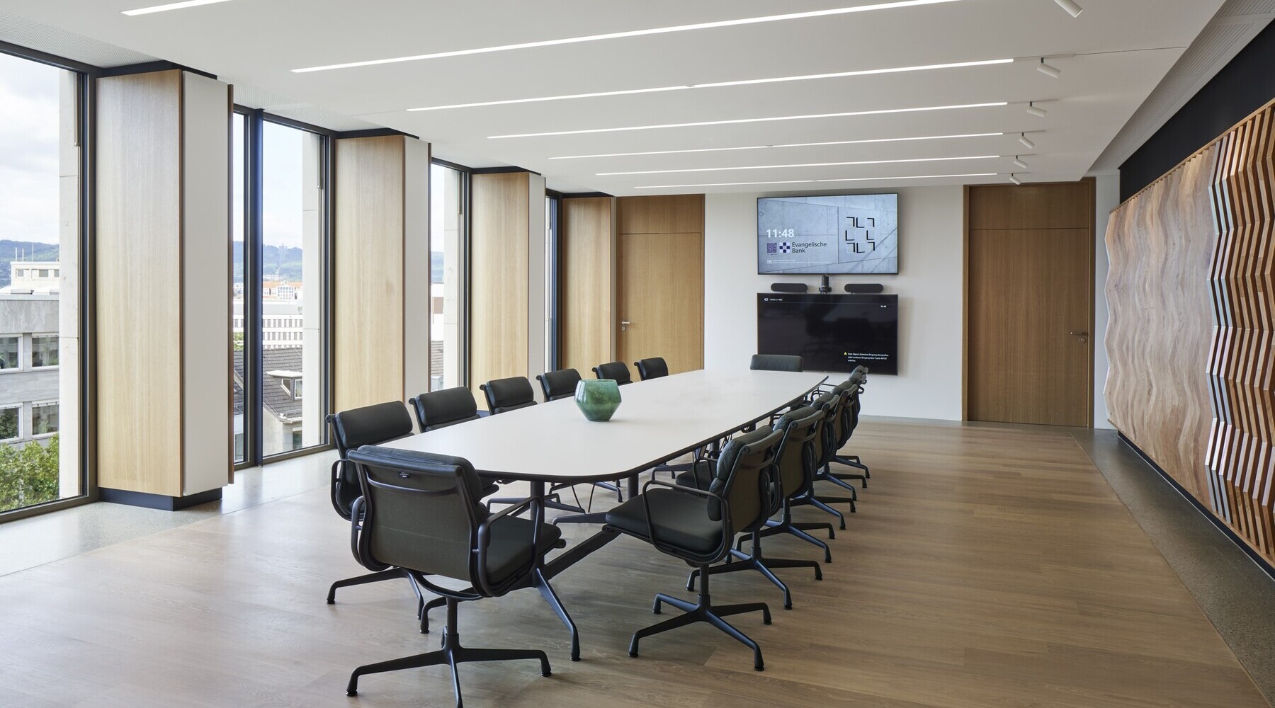 Konferenzraum der EVB mit einem ovalen Meetingtisch und Bürostühlen darum herum in der Mitte, außenherum geben Fenster den Blick nach draußen frei, in der weißen Decke sind lineare Einbauleuchten (PALADIO) sichtbar, zu einer Wandseite sind am Ende der Leuchten noch zylindrische Spots (EVOLUTION SPOT) anschließend eingebaut.