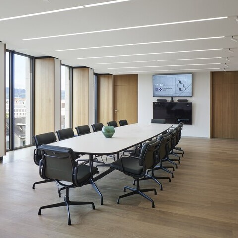 Konferenzraum der EVB mit einem ovalen Meetingtisch und Bürostühlen darum herum in der Mitte, außenherum geben Fenster den Blick nach draußen frei, in der weißen Decke sind lineare Einbauleuchten (PALADIO) sichtbar, zu einer Wandseite sind am Ende der Leuchten noch zylindrische Spots (EVOLUTION SPOT) anschließend eingebaut.