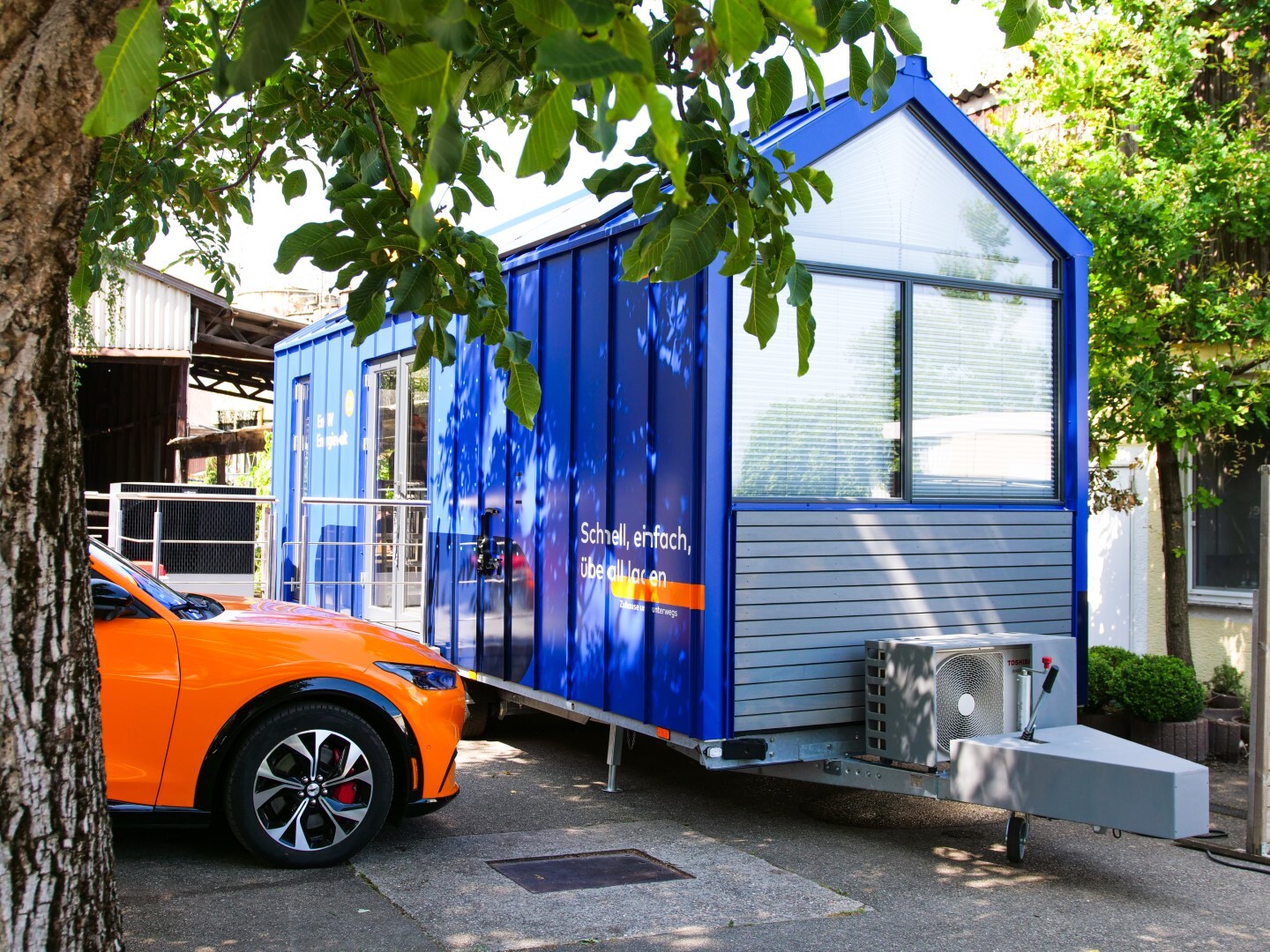 Ein modernes Tiny House in Blau mit großem Fenster, Klimaanlage und Anhängerkupplung.