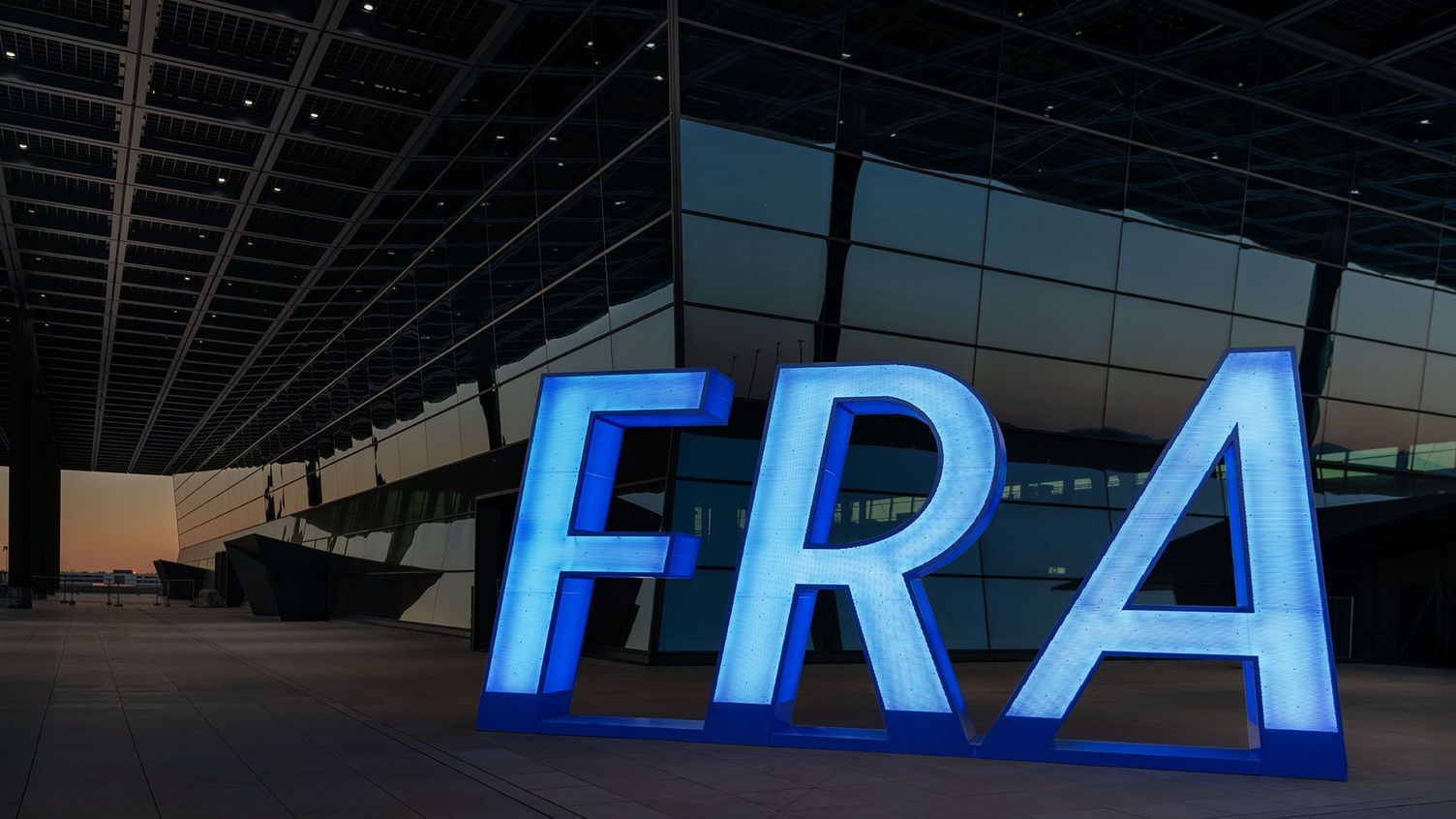 Drei Letter Code FRA für Frankfurter Airport. Außenansicht Frankfurter Flughafen Gebäude mit blau leuchtendem freistehenden Schriftzug FRA an einem Gebäudeeck. | © Fraport