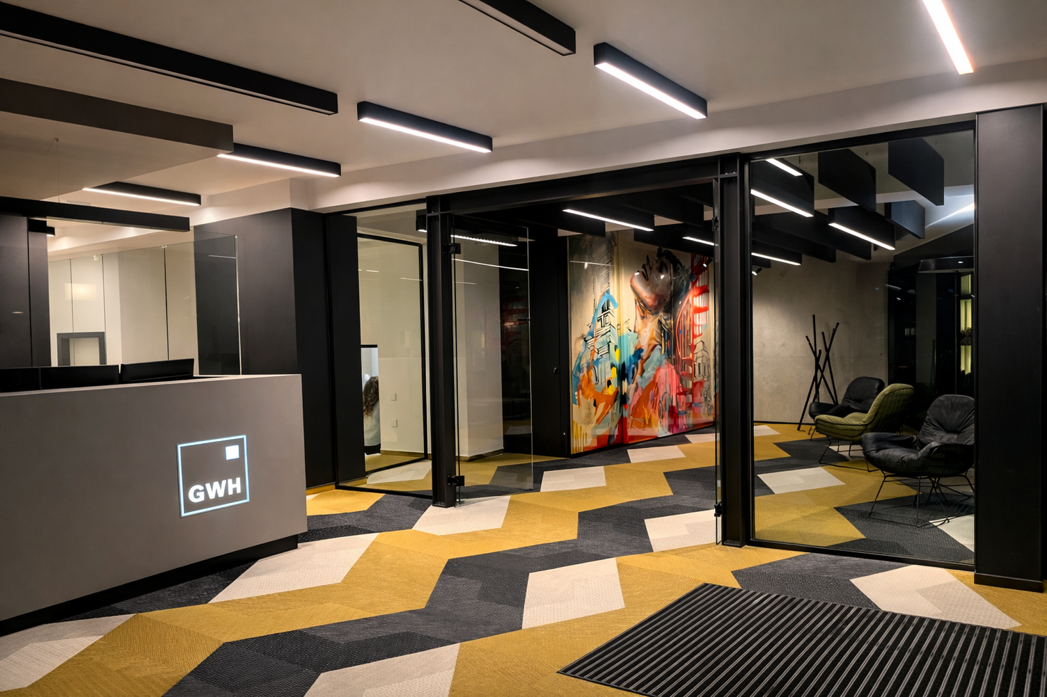 Flur mit Empfangstheke, entlang der Raumdecke lineare Anbauleuchten PALADIO 54 PRISM in zwei unterschiedlichen Längen. In dem Flur liegt wie in der angrenzenden Lounge ein Teppichboden mit gelb, weiß und grauem Zick-Zack-Muster. In der Lounge nebenan stehen zwei moderne Ledersessel in schwarz und grün in der einen Raumecke, daneben ein Kleiderständer aus verschachtelten Holzleisten, darüber auf die komplette Raumlänge sind schwarze Baffeln abgehangen, teils als lineare Akustik-Baffelleuchten ausgestattet mit PALADIO Prism oder PALADIO Tracksystem. Im Track sind mehrere zylindirsche Evolution Spots poistioniert.