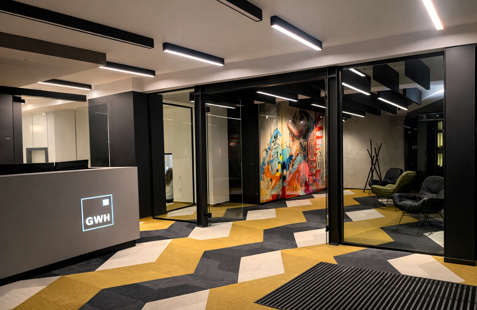 Empfangsbereich mit linearen Anbauleuchten (PALADIO 54 PRISM) Flur mit Empfangstheke, entlang der Raumdecke lineare Anbauleuchten PALADIO 54 PRISM in zwei unterschiedlichen Längen. In dem Flur liegt wie in der angrenzenden Lounge ein Teppichboden mit gelb, weiß und grauem Zick-Zack-Muster. In der Lounge nebenan stehen zwei moderne Ledersessel in schwarz und grün in der einen Raumecke, daneben ein Kleiderständer aus verschachtelten Holzleisten, darüber auf die komplette Raumlänge sind schwarze Baffeln abgehangen, teils als lineare Akustik-Baffelleuchten ausgestattet mit PALADIO Prism oder PALADIO Tracksystem. Im Track sind mehrere zylindirsche Evolution Spots poistioniert.
