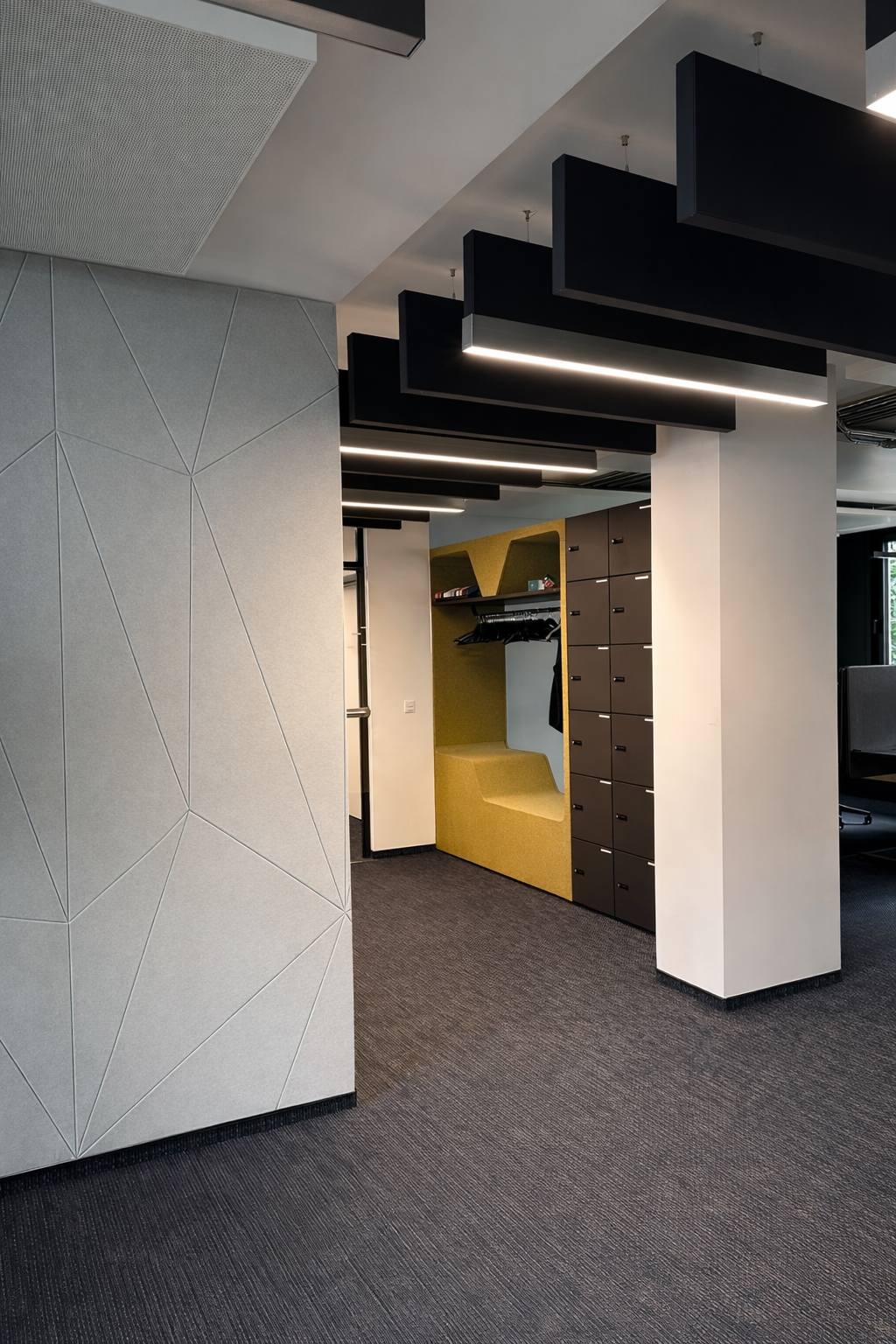Hallway light with baffle lights (black) View from a hallway with dark carpet and light graphic walls into a narrower hallway along which black acoustic baffles are suspended from the ceiling, some of which are acoustic baffles with linear lighting to improve the room acoustics. Baffle lights from the Paladio series by SATTLER.