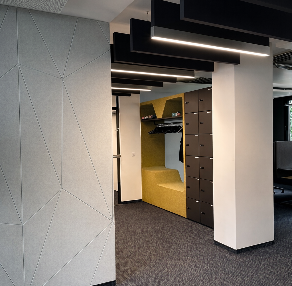 View from a hallway with dark carpet and light graphic walls into a narrower hallway along which black acoustic baffles are suspended from the ceiling, some of which are acoustic baffles with linear lighting to improve the room acoustics. Baffle lights from the Paladio series by SATTLER. 