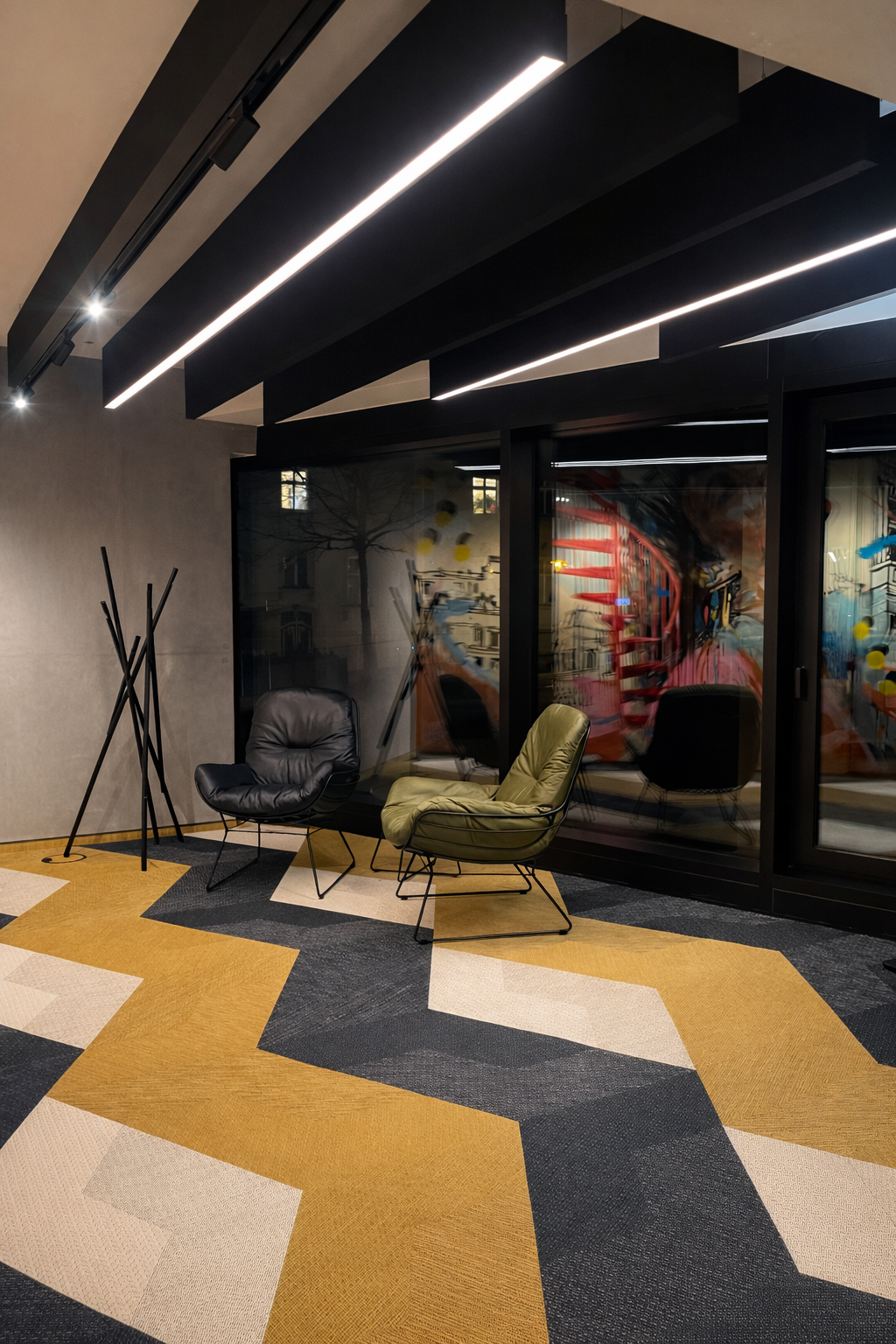 Lounge with graphic carpet featuring a yellow, white and grey zigzag pattern, two modern leather armchairs in black and green in one corner of the room, next to them a coat rack made of interlocking wooden slats, above which black baffles are suspended across the entire length of the room, some of which are equipped with PALADIO Prism or PALADIO track systems as linear acoustic baffle lights. Several cylindrical Evolution spots are positioned in the track.