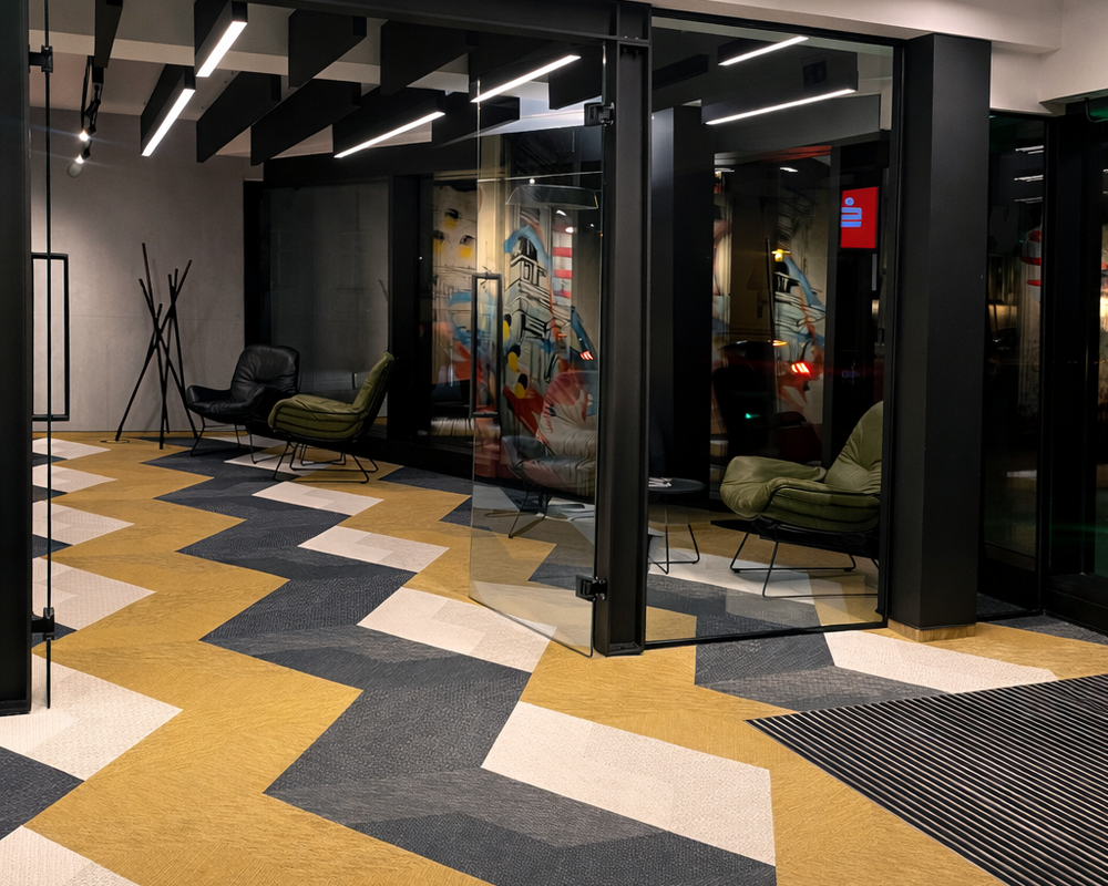 Flur mit lineare Anbauleuchten PALADIO 54 PRISM in zwei unterschiedlichen Längen an der Decke. In dem Flur liegt wie in der angrenzenden Lounge ein Teppichboden mit gelb, weiß und grauem Zick-Zack-Muster. In der Lounge nebenan stehen zwei moderne Ledersessel in schwarz und grün in der einen Raumecke, daneben ein Kleiderständer aus verschachtelten Holzleisten, darüber auf die komplette Raumlänge sind schwarze Baffeln abgehangen, teils als lineare Akustik-Baffelleuchten ausgestattet mit PALADIO Prism oder PALADIO Tracksystem. Im Track sind mehrere zylindirsche Evolution Spots poistioniert.