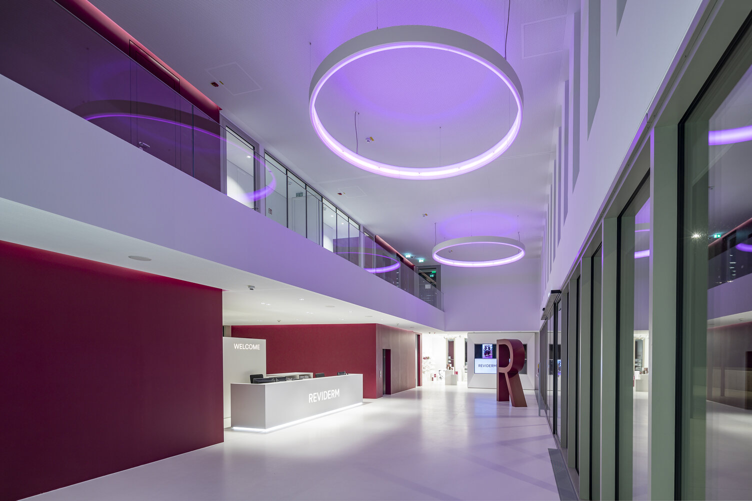Ringleuchte CIRCOLO mit RGB im Eingangsbereich Reviderm | © Michael Heinrich Foyer in Unternehmensfarben von Reviderm in weiß und und einem warmen Rotton. Entlang des Flurs an der Empfangstheke vorbei hängen CIRCOLO Ringleuchten, die in einem lila Ton leuchten. | © Michael Heinrich