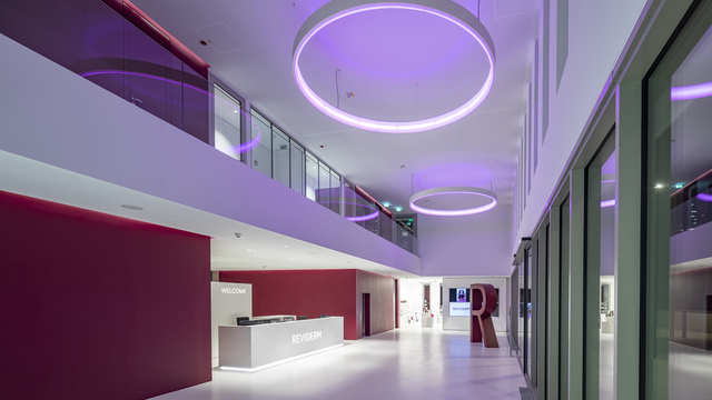 Atrium lighting. Foyer in Reviderm's corporate colours of white and a warm shade of red. Along the corridor past the reception desk hang CIRCOLO ring lights that shine in a purple tone. | © Michael Heinrich | www.licht.studio