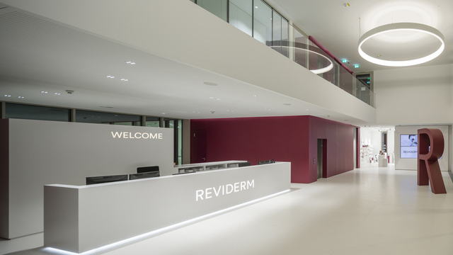 Blick auf Empfangsbereich des Headquarter Reviderm München in Rottönen und weiß gehaltene Wände links im Bild, mittig im Gang mit großen CIRCOLO Ringleuchten in weiß leuchtend. | © Michael Heinrich | www.licht.studio