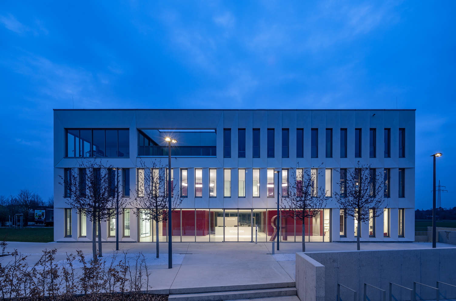 Foyer lighting. Exterior view. View through the exterior façade, consisting of a glass façade on the ground floor and concrete walls interspersed with many long windows on the upper floors. Through the windows, you can see the foyer lighting: two CIRCOLO ring lights in RGB colours at night. | © Michael Heinrich | www.licht.studio