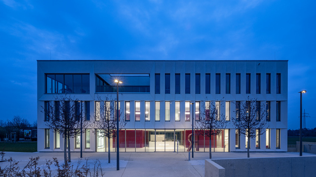 Foyer lighting. Exterior view. View through the exterior façade, consisting of a glass façade on the ground floor and concrete walls interspersed with many long windows on the upper floors. Through the windows, you can see the foyer lighting: two CIRCOLO ring lights in RGB colours at night. | © Michael Heinrich | www.licht.studio