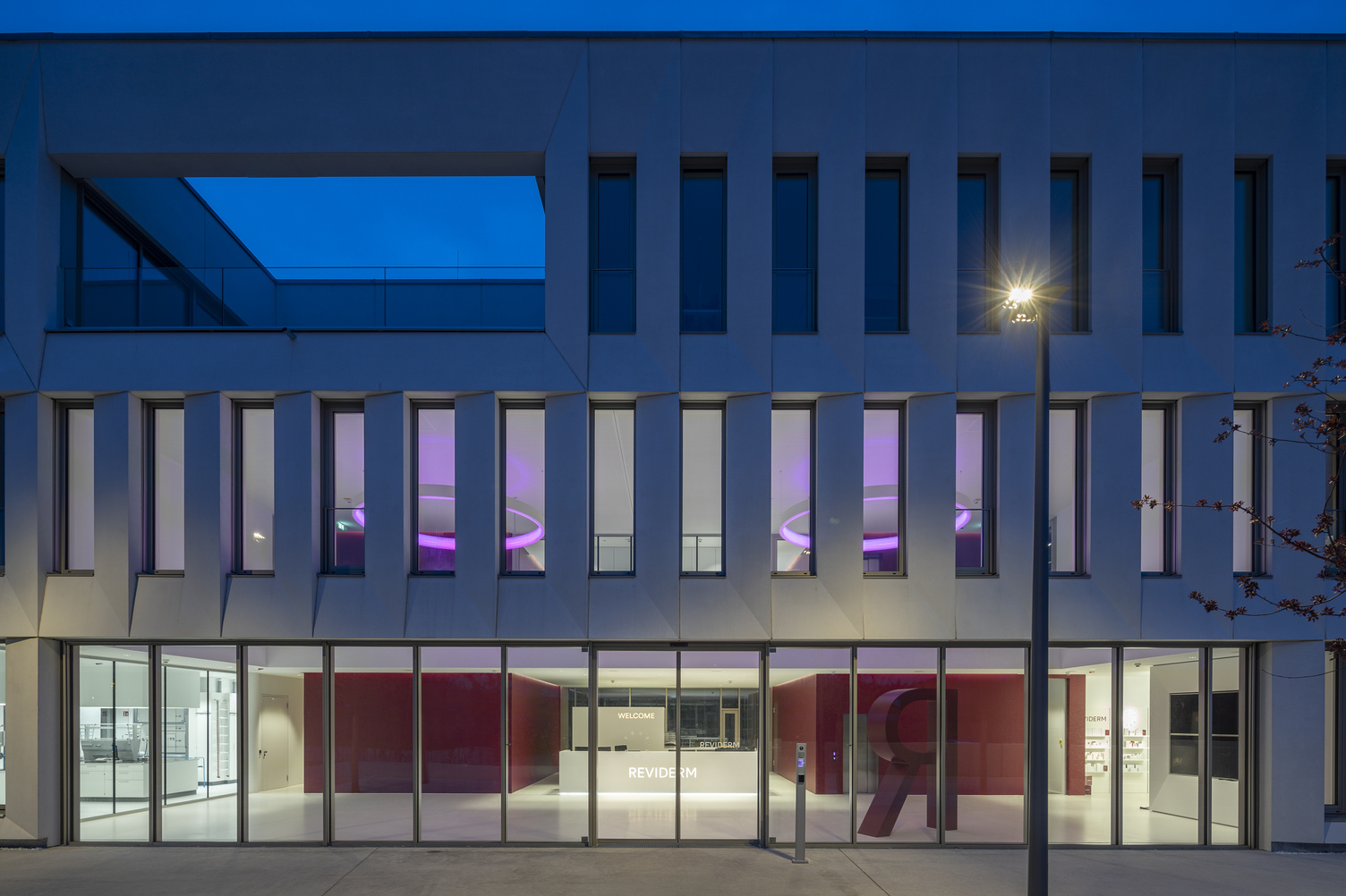 Foyer lighting. Exterior view. View through the exterior façade, consisting of a glass façade on the ground floor and concrete walls interspersed with many long windows on the upper floors. Through the windows, you can see the foyer lighting: two CIRCOLO ring lights in RGB colours at night.