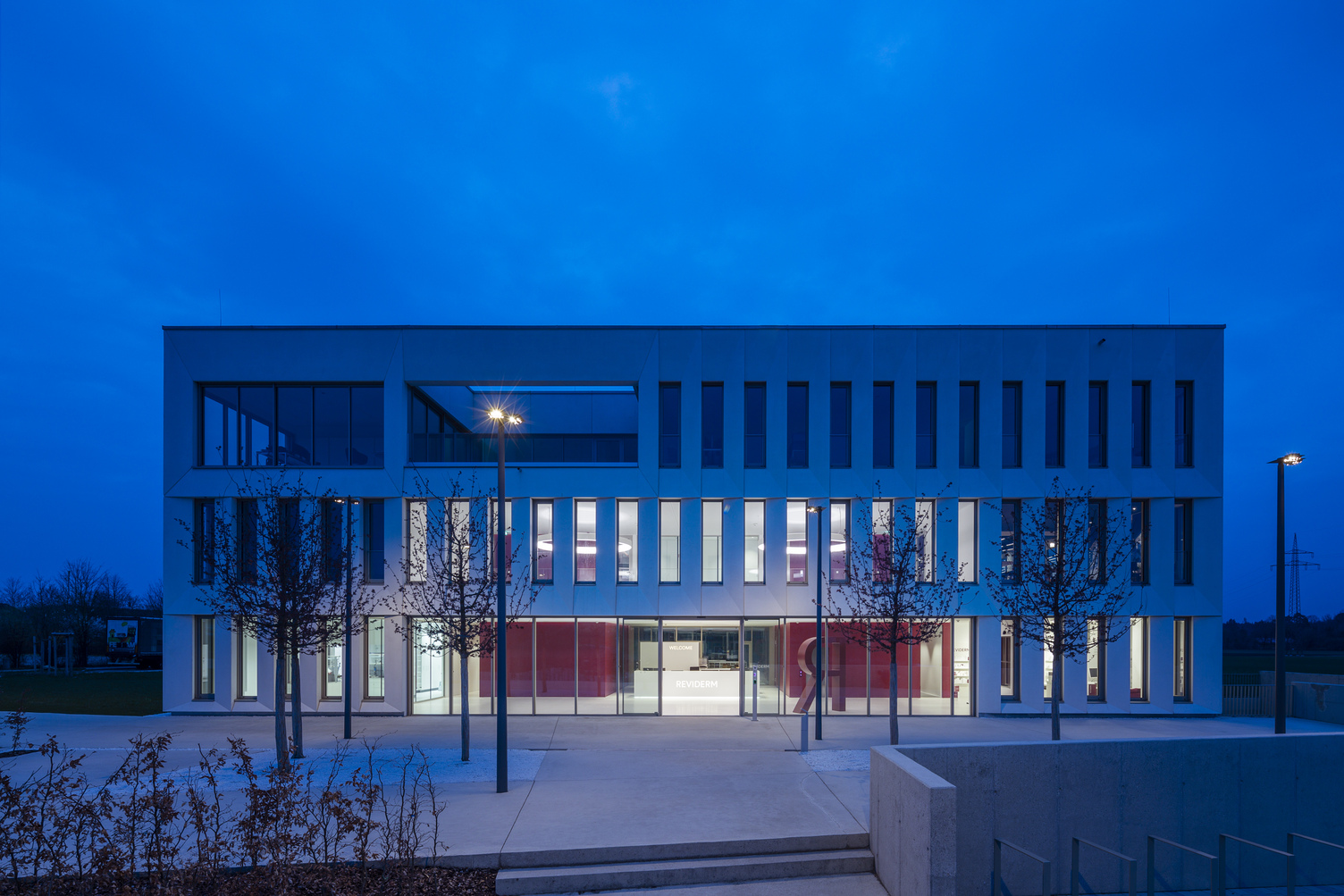 Foyer lighting. Exterior view. View through the exterior façade, consisting of a glass façade on the ground floor and concrete walls interspersed with many long windows on the upper floors. Through the windows, you can see the foyer lighting: two CIRCOLO ring lights in RGB colours at night.