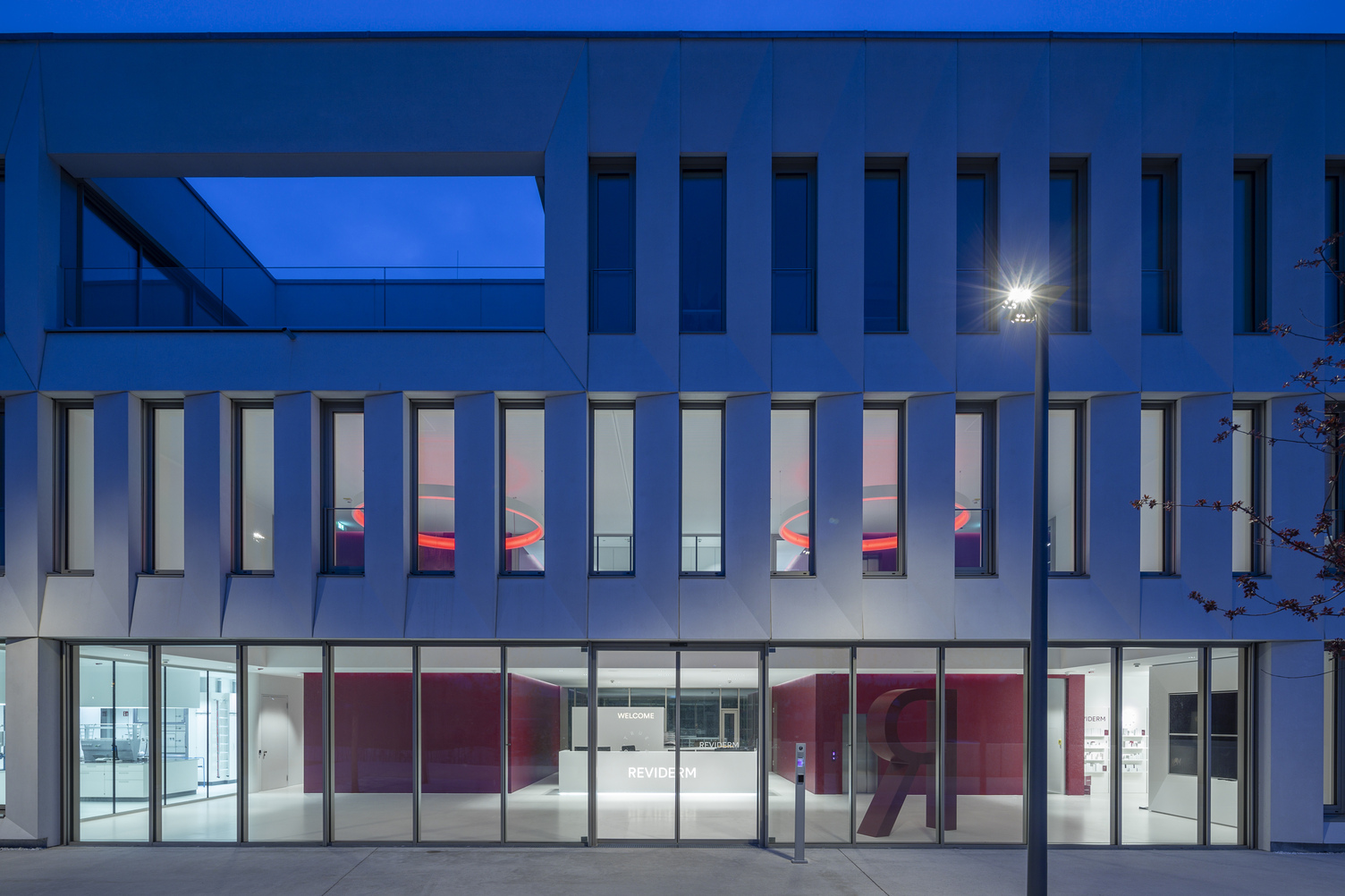 Foyer lighting. Exterior view. View through the exterior façade, consisting of a glass façade on the ground floor and concrete walls interspersed with many long windows on the upper floors. Through the windows, you can see the foyer lighting: two CIRCOLO ring lights in RGB colours at night.