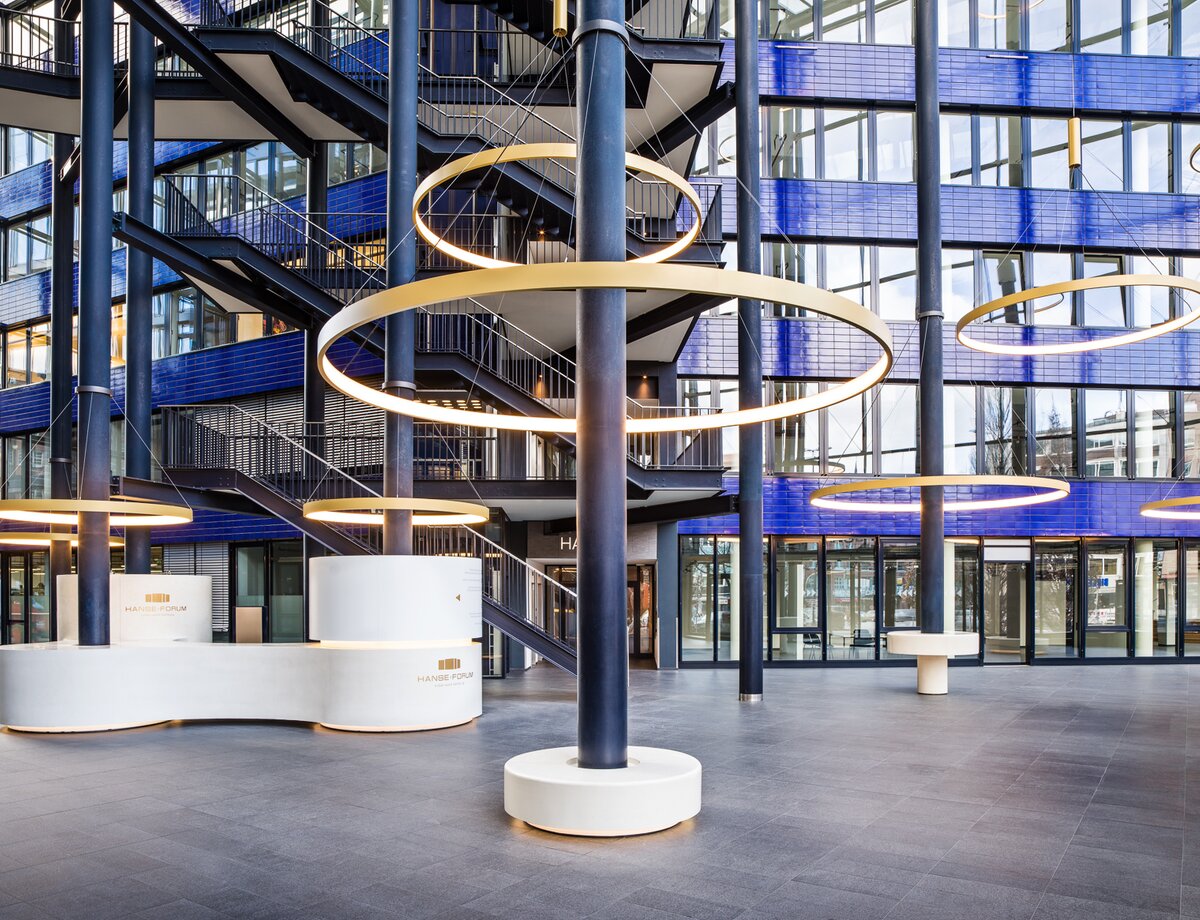CIRCOLO Ringleuchten um Säulen des Atriums Hanseforum Hamburg | © gerrit meier Goldene großformatige Ringleuchten, CIRCOLO I, abgependelt um die Säulen des verglasten Atriums mit blauen Wänden des Hanseforum Hamburg. | © gerrit meier