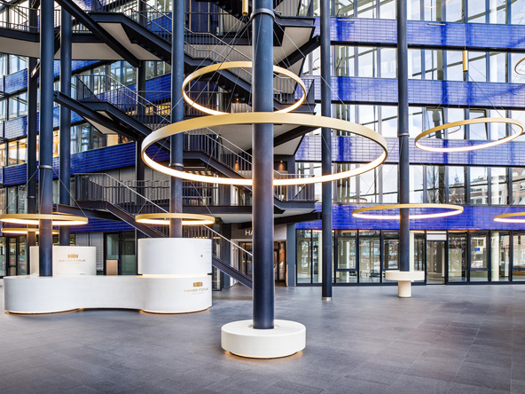 Golden large-format ring lights, CIRCOLO I, suspended around the columns of the glazed atrium with blue walls of the Hanseforum Hamburg. | © gerrit meier