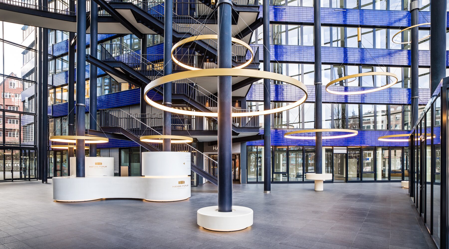 CIRCOLO Ringleuchten um Säulen des Atriums Hanseforum Hamburg | © gerrit meier Goldene großformatige Ringleuchten, CIRCOLO I, abgependelt um die Säulen des verglasten Atriums mit blauen Wänden des Hanseforum Hamburg. | © gerrit meier