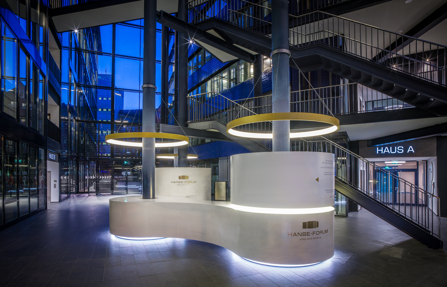 Verbundene s-förmige, weiße, runde Empfangstheke von stayconcrete im Atrium des Hanseforum Hamburg um zwei blaue Säulen, von diesen Säulen ist je eine goldene Ringleuchte CIRCOLO schirmartig abgependelt.
