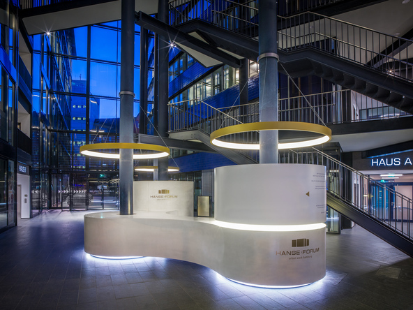 Connected s-shaped, white, round reception desk by stayconcrete in the atrium of the Hanseforum Hamburg around two blue columns, from each of these columns a golden ring light CIRCOLO is suspended like a shade. | © gerrit meier