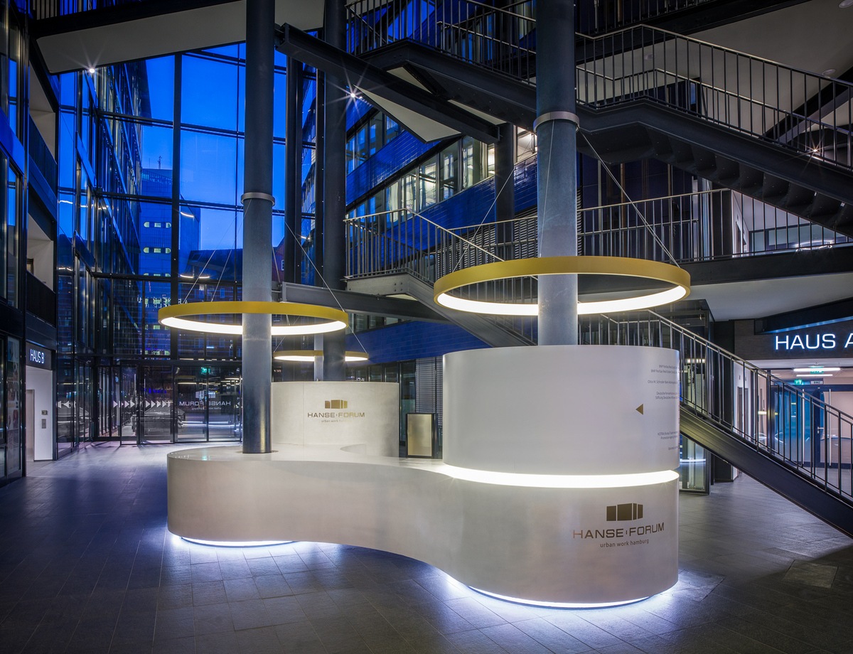 Verbundene s-förmige, weiße, runde Empfangstheke von stayconcrete im Atrium des Hanseforum Hamburg um zwei blaue Säulen, von diesen Säulen ist je eine goldene Ringleuchte CIRCOLO schirmartig abgependelt.