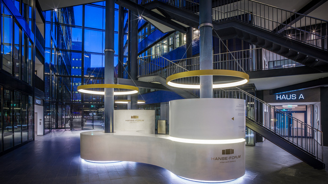 Verbundene s-förmige, weiße, runde Empfangstheke von stayconcrete im Atrium des Hanseforum Hamburg um zwei blaue Säulen, von diesen Säulen ist je eine goldene Ringleuchte CIRCOLO schirmartig abgependelt. | © gerrit meier