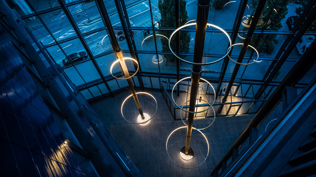 Blick bei Nacht von oben auf das sich dreieckig öffnende Atrium des Hanseforum Hamburg, präsent zeigen sich die blauen Wände und mehrere Säulen in der Raummitte, um welche mehrere Ringlampen CIRCOLO abgehangen sind und den Raum erleuchten. | © gerrit meier