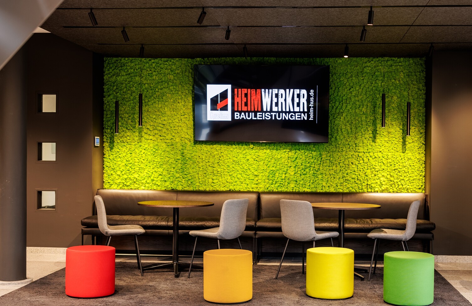 Colourful waiting lounge at Heim Infrastrukturbau with a green wall in front of which cylindrical pendant luminaires (TIA) are suspended and a light track system with spotlights on the ceiling. | © Tobias Fröhner Photography