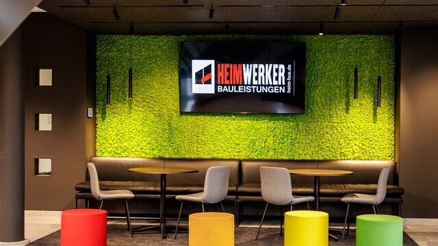 Colourful waiting lounge at Heim Infrastrukturbau with a green wall in front of which cylindrical pendant luminaires (TIA) are suspended and a light track system with spotlights on the ceiling. | © Tobias Fröhner Photography