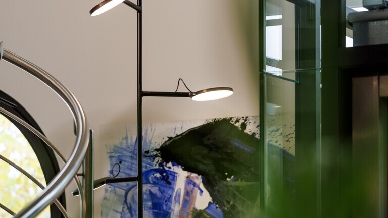 Vertically tilted black pendant luminaire with light heads, AVVENI VERTICAL 3, in front of a colourful mural, to the left of it a curved metal stair railing. | © Tobias Fröhner Photography