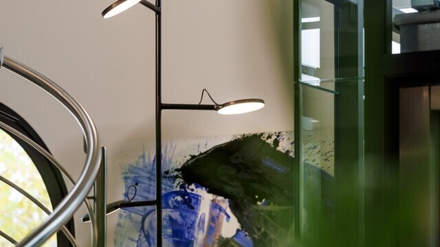 Vertically tilted black pendant luminaire with light heads, AVVENI VERTICAL 3, in front of a colourful mural, to the left of it a curved metal stair railing. | © Tobias Fröhner Photography
