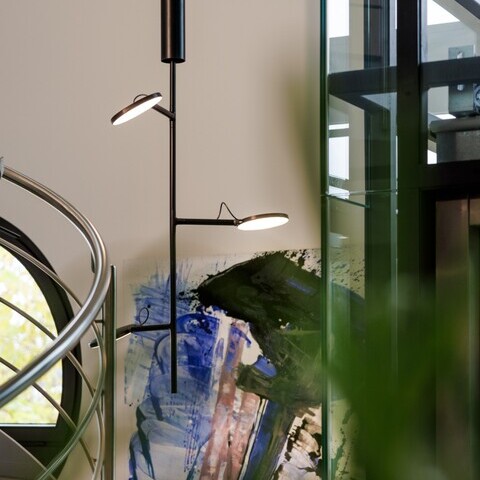 Vertikal verzweigte schwarze Pendelleuchte mit Leuchtköpfen, AVVENI VERTICAL 3, vor einem bunten Wandbild, links daneben gebogenes Metalltreppengeländer.