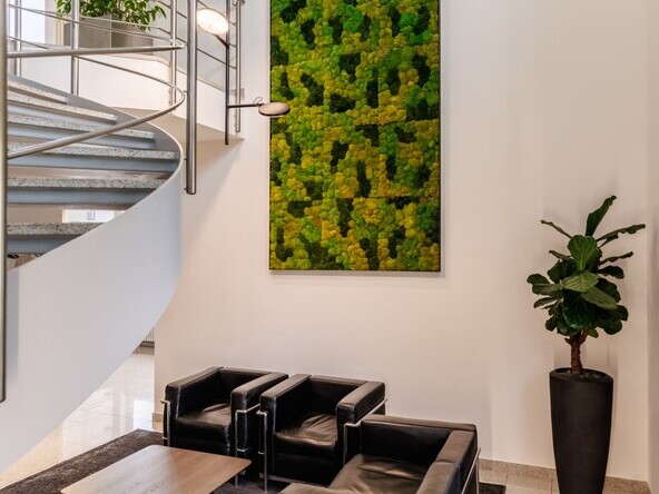 Vertically branched black pendant lamp with round light heads, AVVENI VERTICAL 4, which shines in different directions – downwards and to different sides. Suspended above a lounge area with two black leather armchairs and two colourful stools, in front of a moss wall mural, with a curved metal stair railing to the left. | © Tobias Fröhner Photography