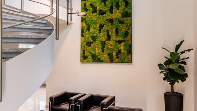 Vertically branched black pendant lamp with round light heads, AVVENI VERTICAL 4, which shines in different directions – downwards and to different sides. Suspended above a lounge area with two black leather armchairs and two colourful stools, in front of a moss wall mural, with a curved metal stair railing to the left. | © Tobias Fröhner Photography