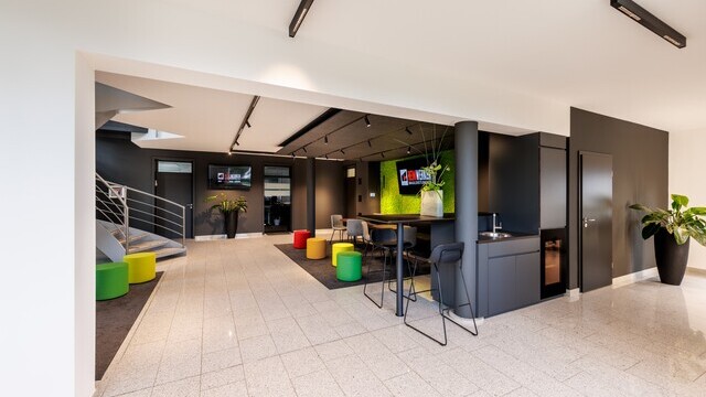 Room with a wide, bright hallway that runs from the right, continues centrally across the corner and past a dark lounge area with colourful stools. Several black linear ceiling-mounted luminaires, some with individual cylindrical spots, PALADIO LENS with EVOLUTION SPOTS, can be seen above the hallway. | © Tobias Fröhner Photography