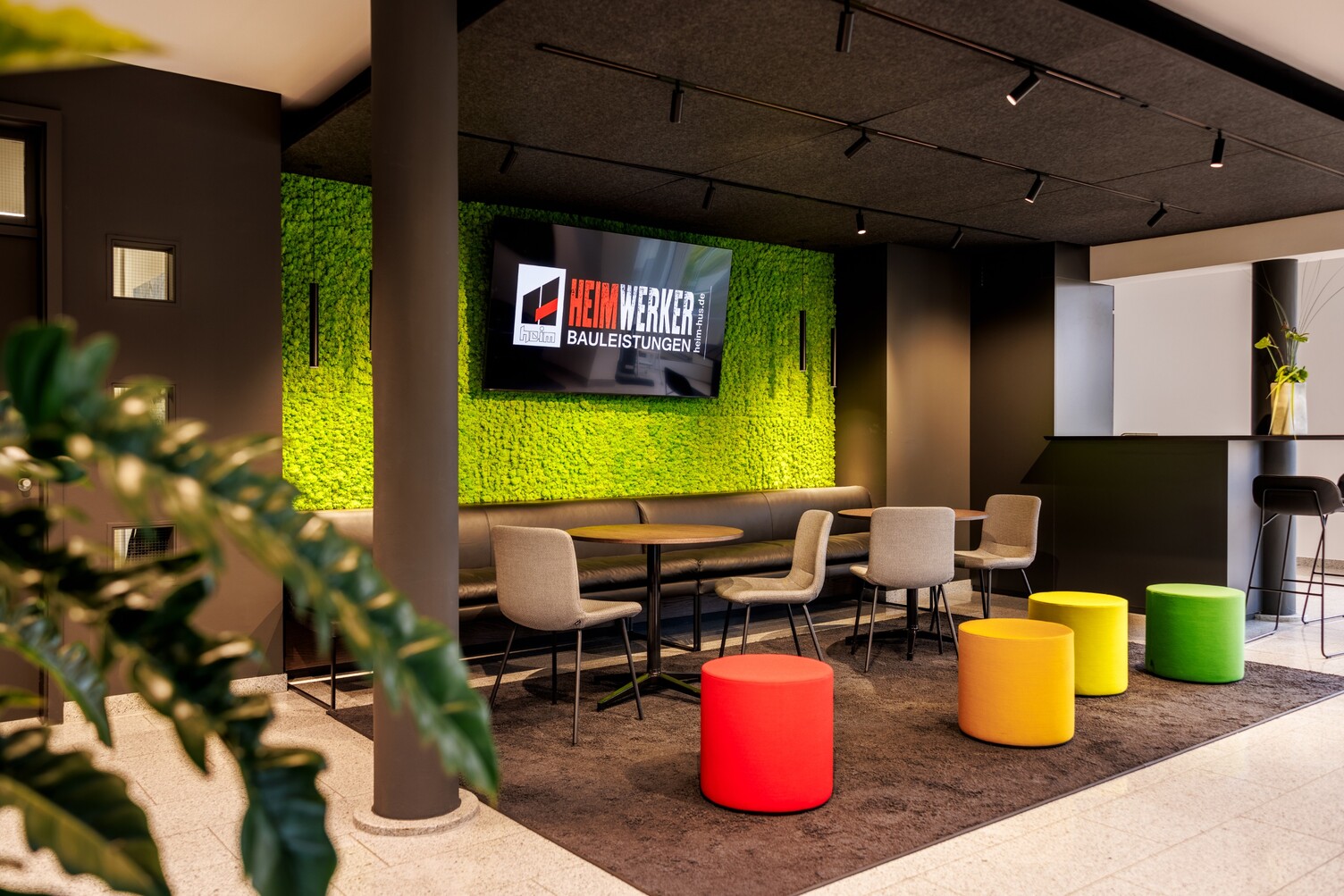 Zurückhaltende, flexible Loungebeleuchtung Blick auf einen Raum im Raum. Im Fokus ein dunkel gehaltener Loungebereich mit einer knallig grünen Rückwand mit mittig positioniertem Bildschirm, 4 bunten Hockern im Vordergrund und beleuchtet über 3 Einbaustromschienen mit zylindrischen EVOLUTION SPOTs, die in verschiedene Richtungen zeigen & zylindrische Pendelleuchten, TIAs, nahe an der grünen Wand, links und rechts neben Bildschirm.