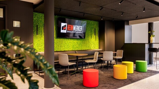 View of a room within a room. The focus is on a dark lounge area with a bright green back wall and a centrally positioned screen, four colourful stools in the foreground and lighting provided by three recessed power rails with cylindrical EVOLUTION SPOTs pointing in different directions and cylindrical pendant lights, TIAs, close to the green wall, to the left and right of the screen. | © Tobias Fröhner Photography