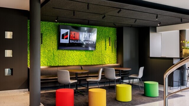 The image shows a bright corridor with a black linear luminaire, PALADIO, running parallel above it. In the background is a lounge area with a dark ceiling, bright green back wall and colourful stools in red, orange, yellow and green. | © Tobias Fröhner Photography