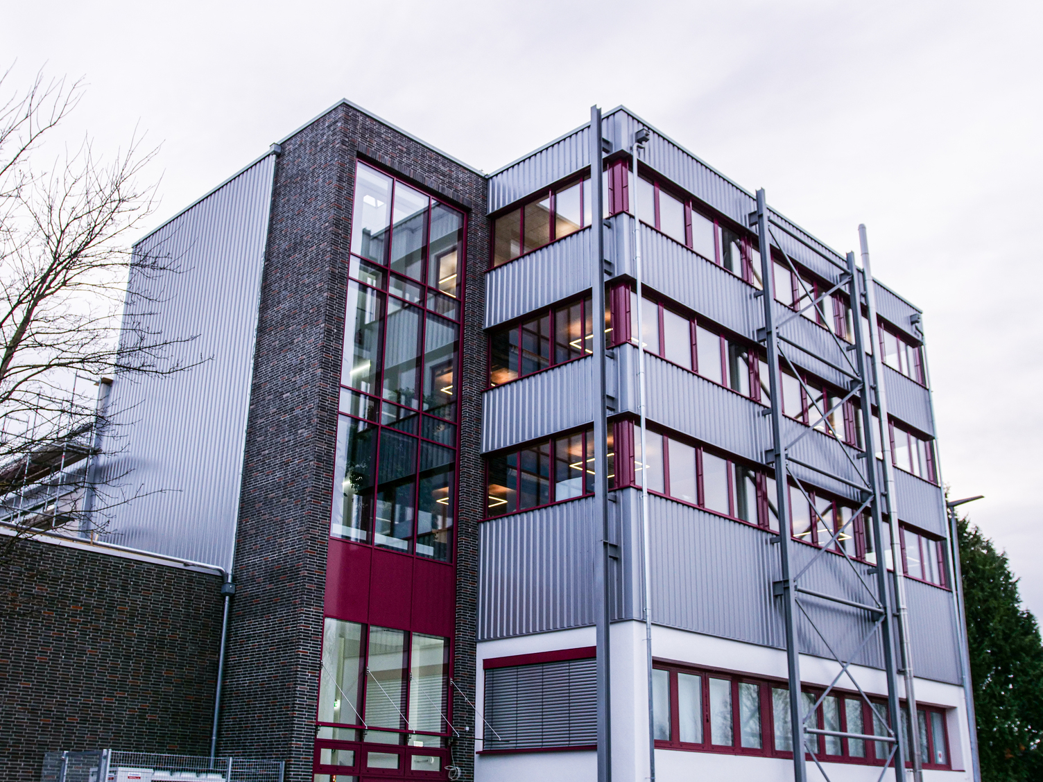Moderne Bürofassade über Eck mit silbrig glänzender Metallverkleidung und bordeauxroten Fensterbändern, dahinter beleuchtete Innenräume