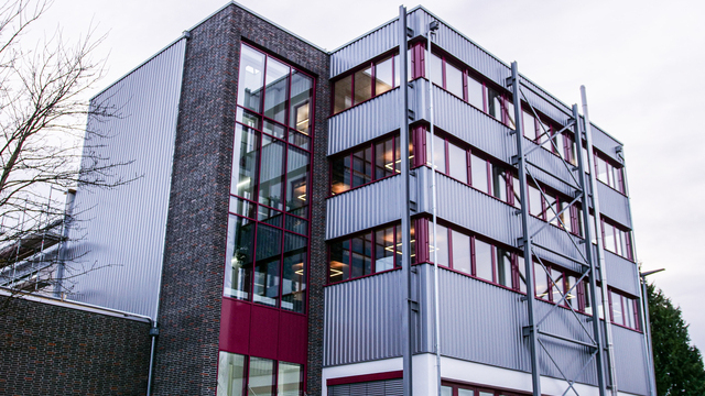 Moderne Bürofassade über Eck mit silbrig glänzender Metallverkleidung und bordeauxroten Fensterbändern, dahinter beleuchtete Innenräume
