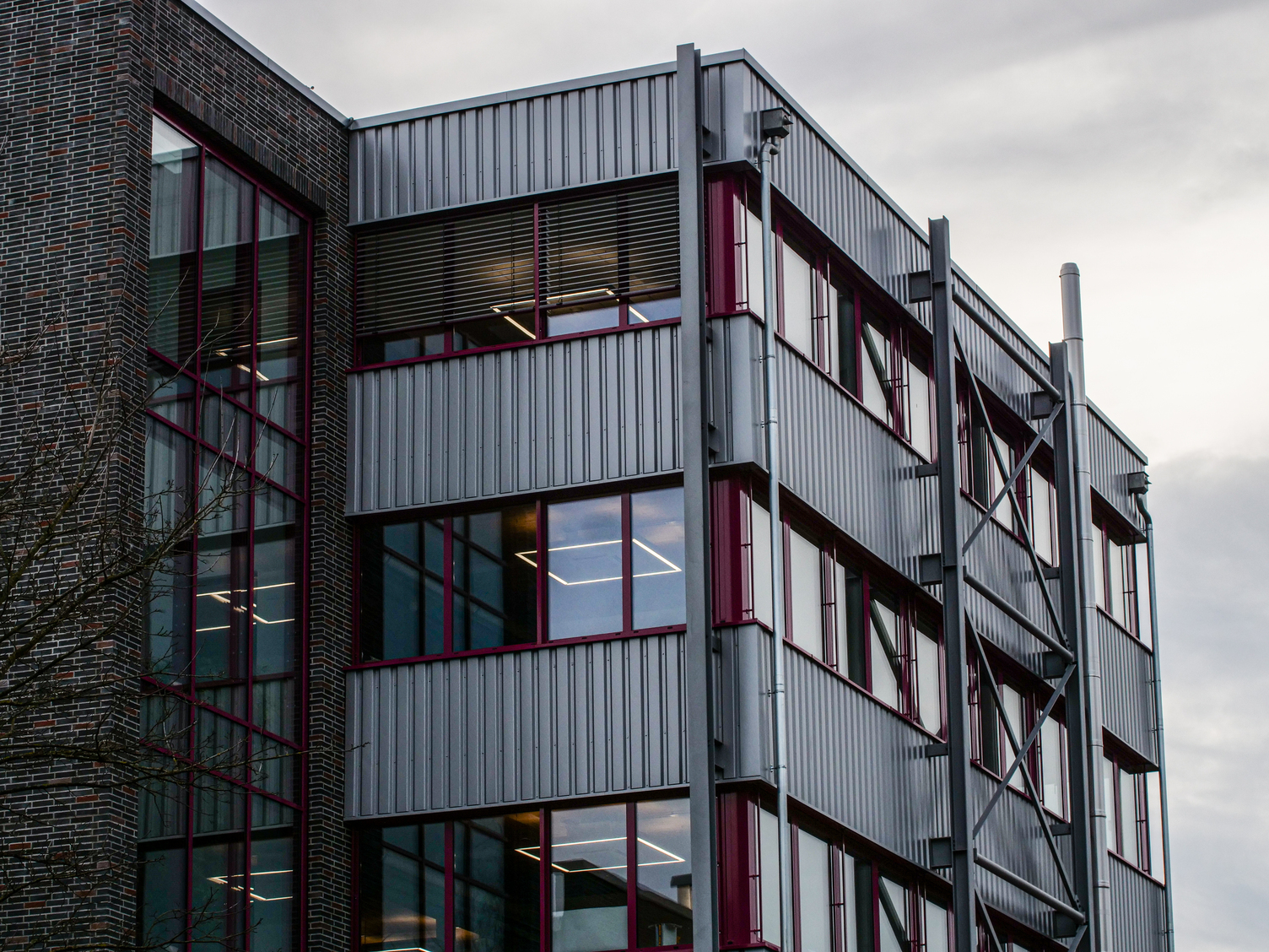 Aufsicht auf die geriffelte Metallfassade eines Firmengebäudes mit bordeauxroten Fensterrahmen; in den Räumen sind lineare Lichtlinien erkennbar
