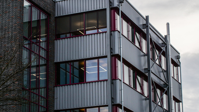 Aufsicht auf die geriffelte Metallfassade eines Firmengebäudes mit bordeauxroten Fensterrahmen; in den Räumen sind lineare Lichtlinien erkennbar