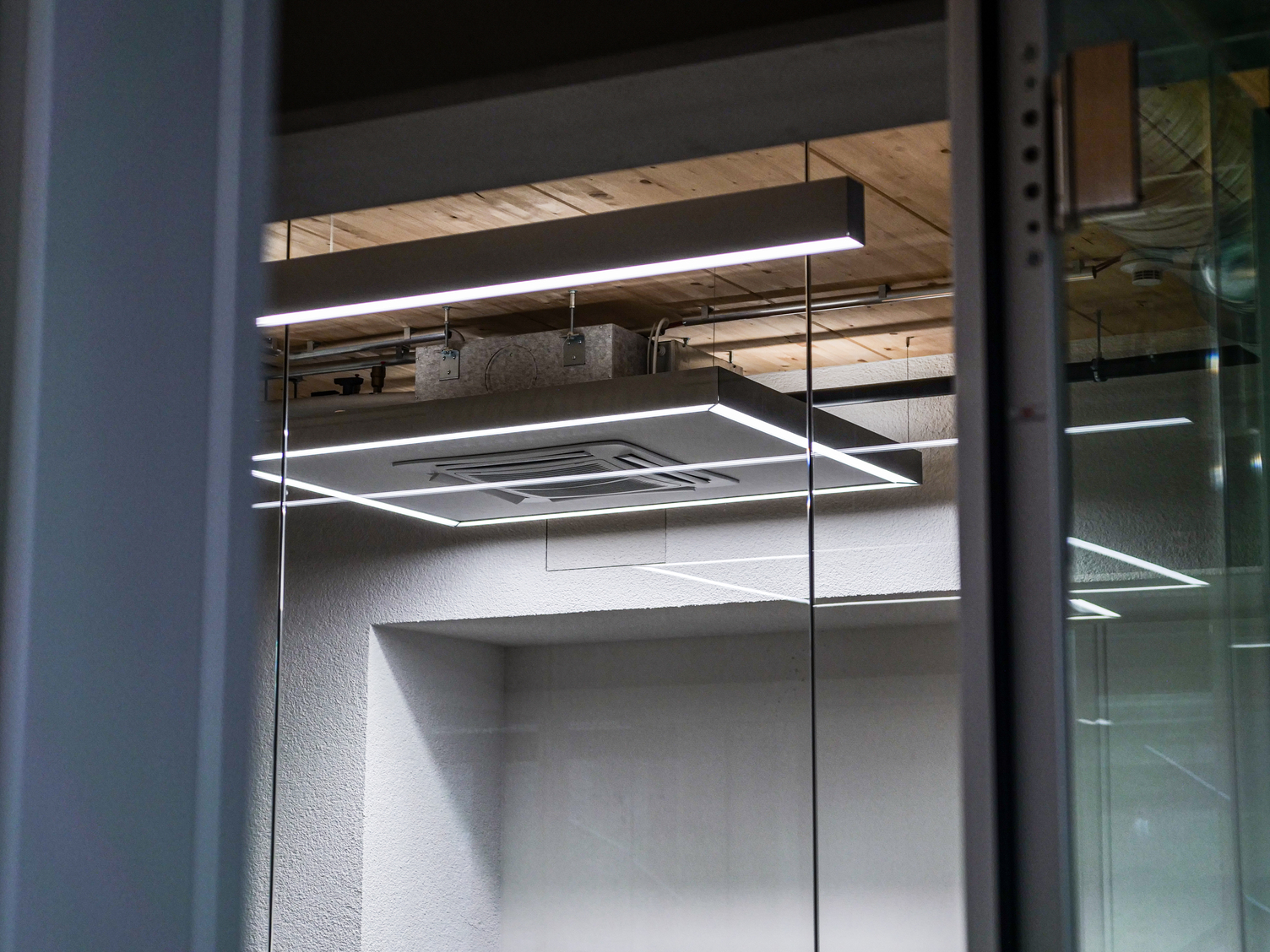Quadratische Ringleuchte und darüber eine Linearleuchte an einer Industriedecke mit Holzelementen, im Hintergrund ein verglaster Aufzug