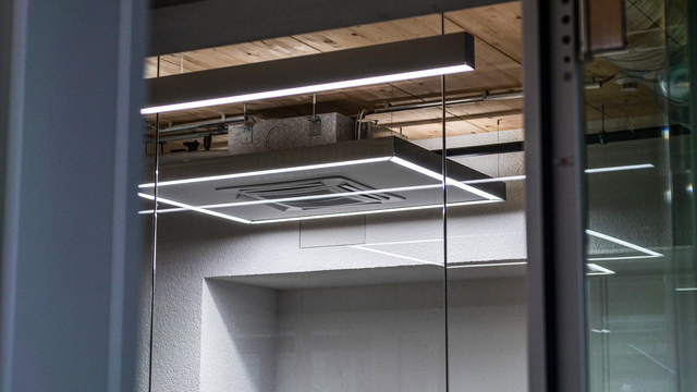 Quadratische Ringleuchte und darüber eine Linearleuchte an einer Industriedecke mit Holzelementen, im Hintergrund ein verglaster Aufzug