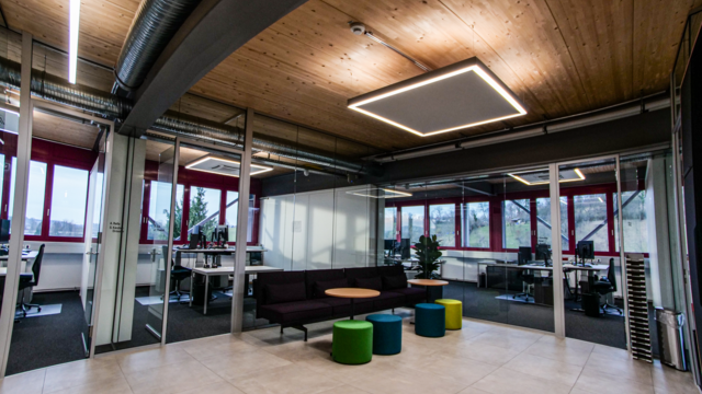 Loungebereich mit quadratischer Ringleuchte und Linearleuchten an einer hölzernen Tonnendecke, Glastrennwänden, dunklem Sofa und bunten Sitzhockern