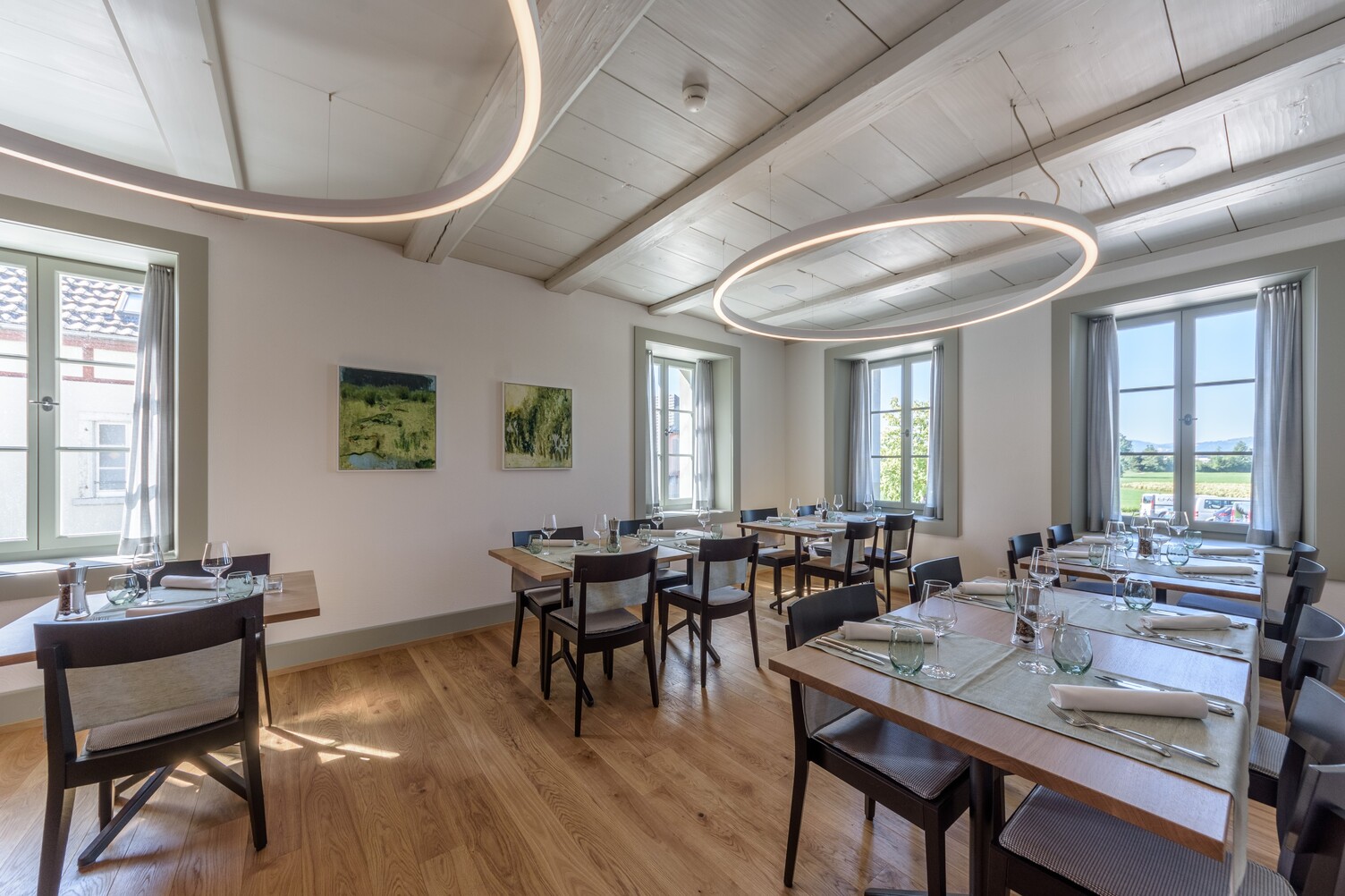 Elegant and cosy restaurant room in the Hirschenbad Langenthal, invitingly laid tables, white wooden ceiling from which large, narrow, directly illuminated white ring lights (DUETTO) are suspended.