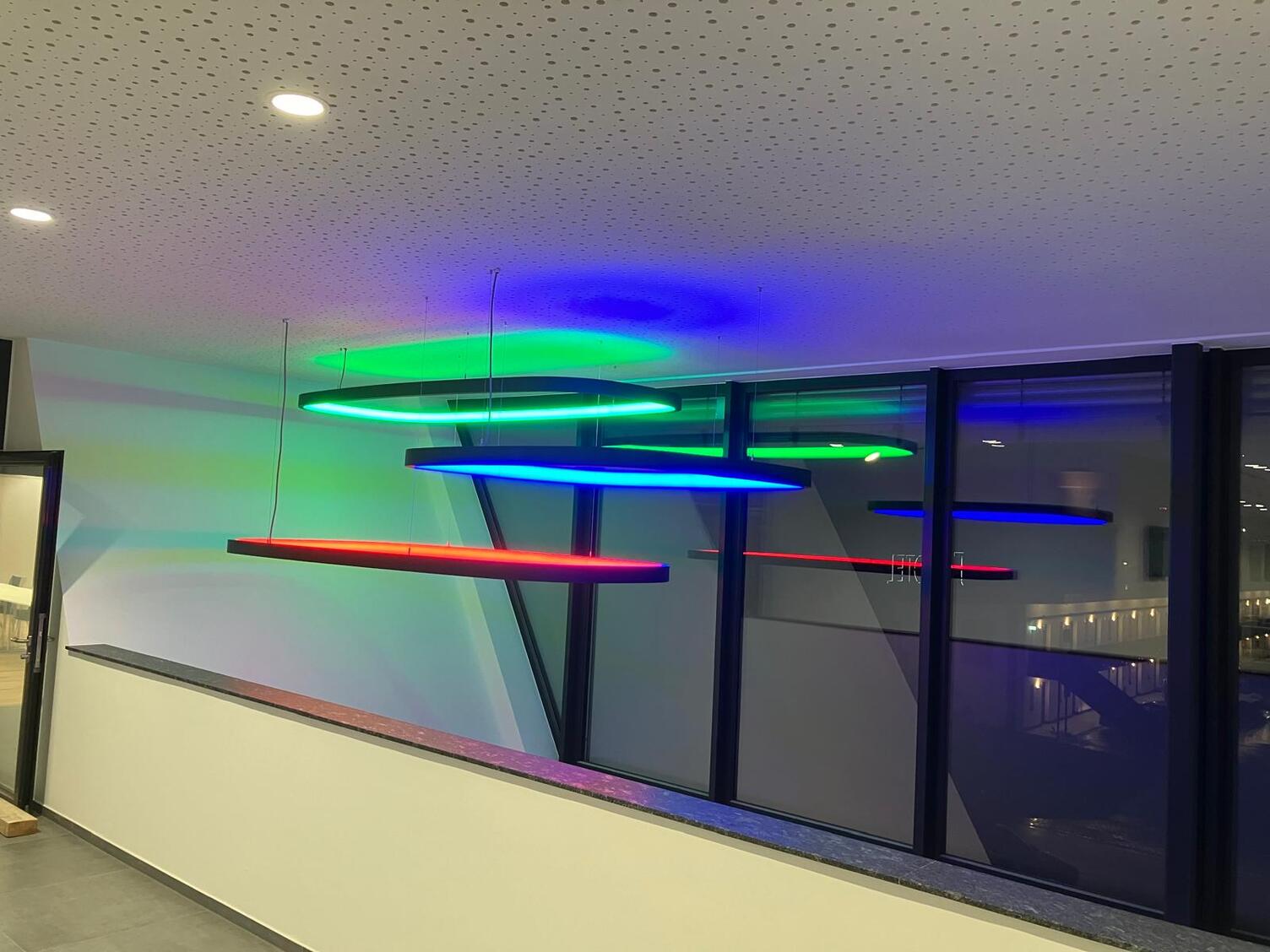 SOFT SQUARE mit RGB-Licht - Rechteckige Ringpendelleuchten im Treppenhaus Blick von einem Büro Balkonflur der KSK-Bau Freiburg auf mehrere rechteckige großformatige Ringleuchten SOFT SQUARE mit schwarzem Profil, die in den verschiedenen RGB-Farben leuchten und in verschiedenen Höhen von der Decke abgehangen sind.